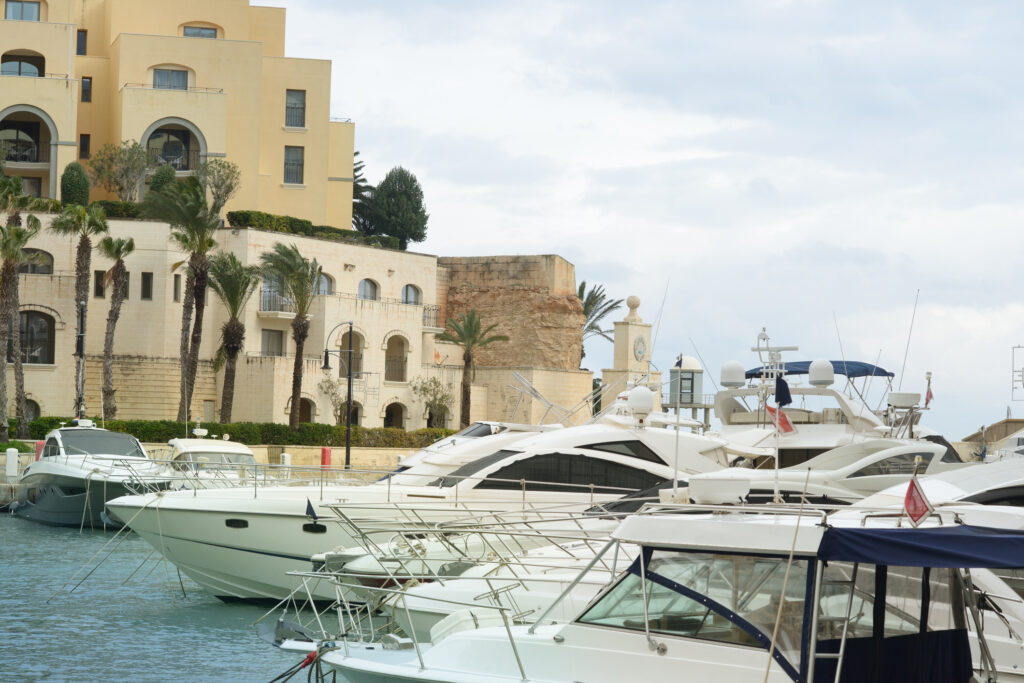 Luxury yachts moored at a marina in Portomaso, Malta, with historical architecture and palm trees in the background